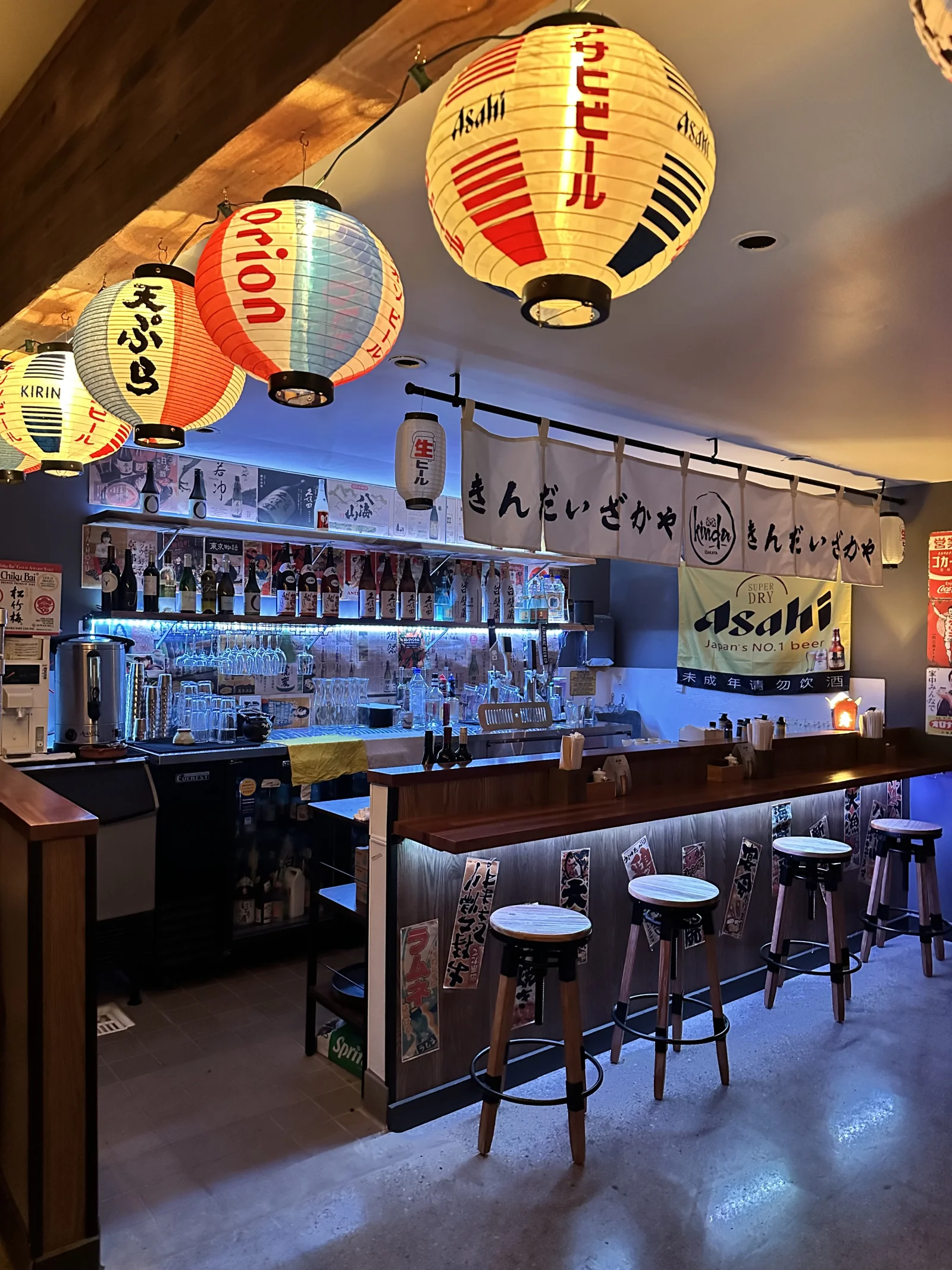 White and red paper lanterns above Kinda’s wooden bar, blue-lit sake shelves and black bar stools.